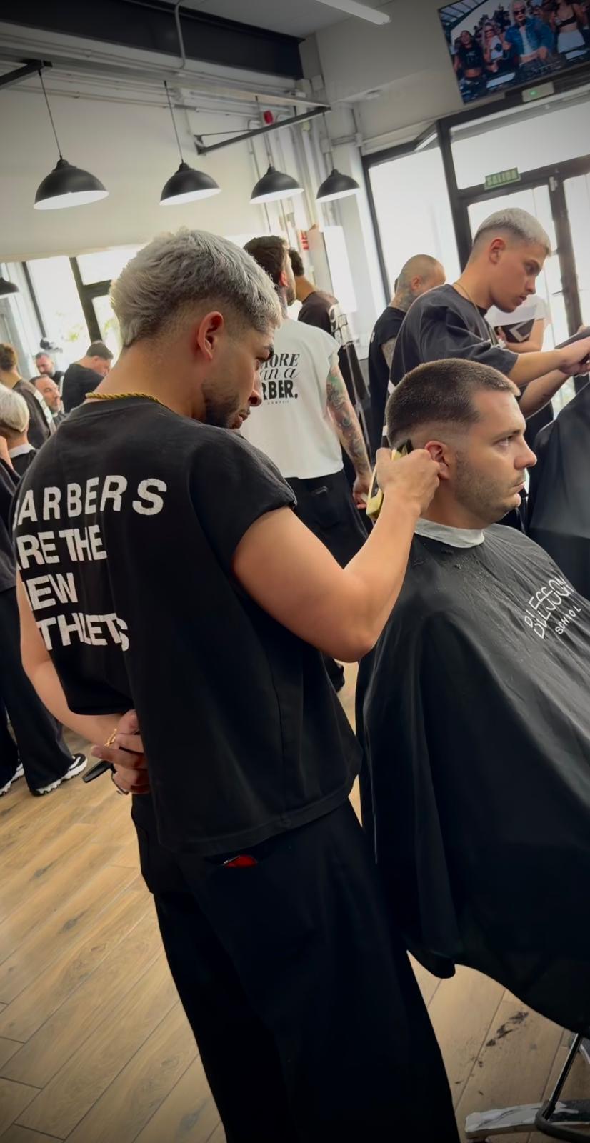 Un barbero pasa la máquina alrededor de la oreja de un cliente sentado mientras se realizan otros cortes en la sala.